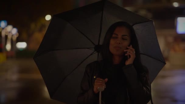 夜の雨の中で携帯電話の外で傘を持っている若い女性 — ストック動画