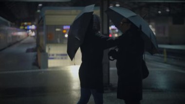 Resmi giyinmiş bir erkek ve bir kadın yağmurda şemsiyelerin altında dikiliyorlar, etrafı karanlıkla çevrili. Elektrikli mavi şemsiyeler binaların cam cephesine karşı kontrasttır.