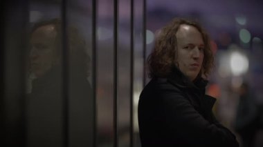 Gece yarısı, uzun saçlı bir adam cam bir duvarın önündeki karanlık bir odada duruyor. Ahşap kaplama gizemli atmosfere ekleniyor.