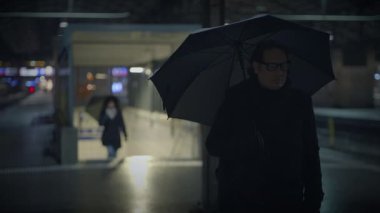 Yağmurun altında duran bir adam yağmurdan korunmak için şemsiye tutuyor. Karanlık, ıslak asfalttan yansıyan otomotiv ışıklarıyla aydınlanır.