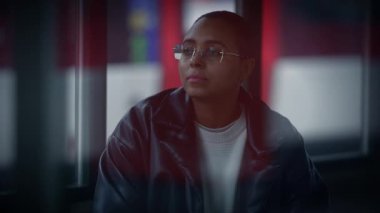 Gözlüklü ve deri ceketli bir kadın karanlıkta pencerenin kenarında oturur. Müzik ve ses ekipmanları ile bir eğlence etkinliği ipuçları dışında bir tabela üzerindeki morumsu yazı tipi