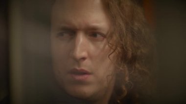 Uzun kıvırcık saçlı bir adam tren istasyonunda insanları seyrediyor.