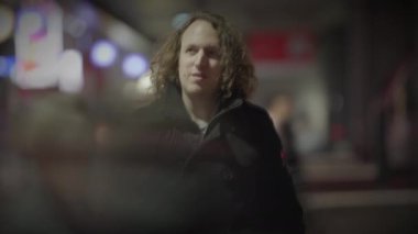 Uzun kıvırcık saçlı bir adam tren istasyonunda insanları seyrediyor.