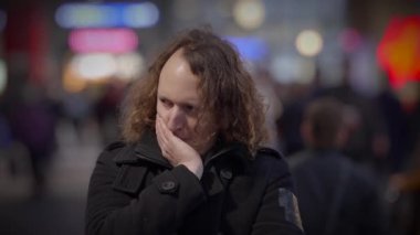 Uzun kıvırcık, mavi saçlı bir adam karanlıkta gülümsüyor. Kalabalık bir caddedeki eğlenceli bir etkinlikte ağzını eliyle kapatıyor.