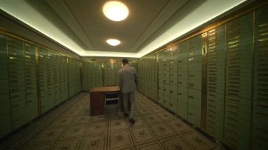 İyi giyimli bir adam binaların koridorunda elinde bir çantayla geziniyor. Döşeme sert ahşaptan, tavan yüksek ve duvarlar bileşik malzemelerden yapılmış.