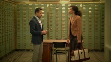 Takım elbiseli bir adam banka kasasında bir kadınla ahşap döşeme seçeneklerini tartışıyor. Ahşap leke ve resmi bir etkinlik masası hakkında fikir paylaşıyorlar.