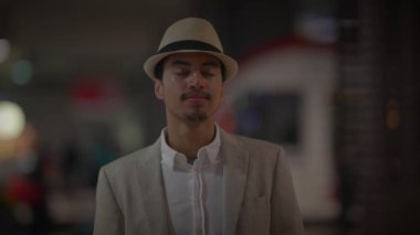 Şık bir genç adam şapka ve ceket giyiyor, gece hayatı ortamında gözleri kapalı duruyor..