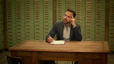 Eski bir ofiste dosya dolapları olan ahşap bir masaya notlar yazarken derin düşüncelere dalmış bir işadamı..