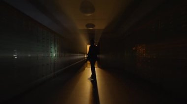 Gölgeli bir şahıs karanlık bir koridorda parıldayan bir kapıya doğru yürüyor ve şüpheli bir atmosfer yaratıyor..