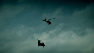 İki askeri helikopter gökyüzünde havacılık manevraları yapıyor. Bulutlar, savunma ve taktik operasyonlar gösteriyor.