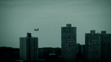 Gün batımında, dramatik bir gökyüzüne karşı siluetlenmiş askeri helikopterler şehir manzarası üzerinde uçarken görülürler.
