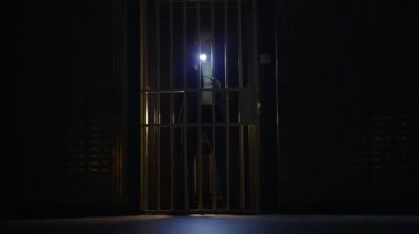 Loş bir odada parmaklıkların arkasında duran el fenerli bir figür gizem ve gerilim hissi uyandırıyor.