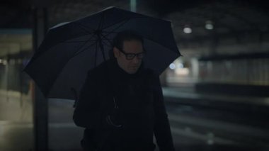 Gece loş bir tren istasyonunda şemsiyeyle yürüyen bir adam kasvetli ve gizemli bir atmosfer yaratıyor.