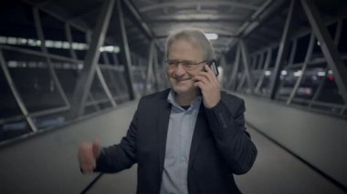 Takım elbiseli bir işadamı gece vakti modern kapalı bir yürüyüş yolunda yürürken telefonuyla mutlu bir şekilde konuşuyor..
