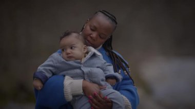 Bir anne bebeğini sevgiyle kucağına alır, ikisi de sıcacık giyinir, doğal arka planı olan açık bir ortamda..