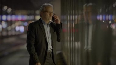 Takım elbiseli yaşlı bir işadamı gece şehirde arka planda bulanık ışıklar olan bir akıllı telefon kullanıyor.