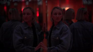 Kot ceketli bir kadın kırmızı ışıklı bir aynanın karşısında duruyor, yansıtıcı ve düşünceli bir atmosfer yaratıyor..