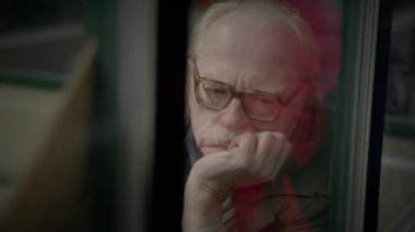 Gözlüklü yaşlı adam pencereden dışarı bakar düşüncelere dalmış, hayatı ve deneyimleri yansıtır.