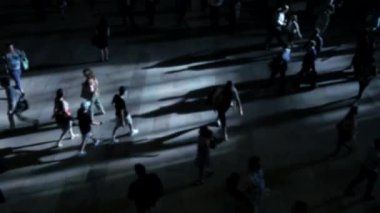 Karanlık bir sokakta yürüyen, çarpıcı ışıklarla aydınlanan, uzun gölgeler yaratan insanların yukarıdan görünüşü..