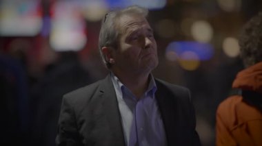 Takım elbiseli, düşünceli, orta yaşlı bir adam şehrin bulanık ışıklarının arka planına dikkatlice bakıyor.