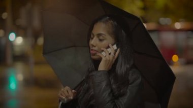 Geceleri elinde şemsiyeyle cep telefonuyla konuşan koyu renk saçlı bir kadın. Arka planda bulanık şehir ışıkları var.