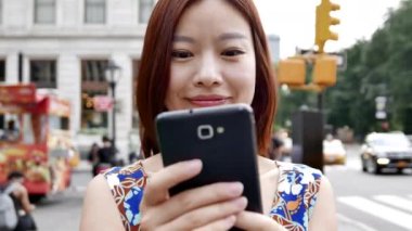 Kalabalık bir şehirde mutlu bir kadın karşıdan karşıya geçerken akıllı telefon kullanır, modern şehir bağlantılarından keyif alır.