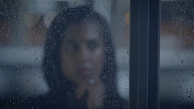 Yağmurlu bir pencerenin arkasında yüzü bulanık, düşünceli bir kadın, yansıma ve melankoli hissi uyandırıyor..