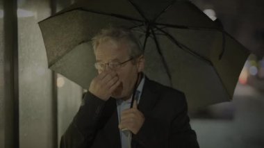 Şemsiyenin altında saklanan yaşlı bir adam, yağmurlu bir ortamda burnunu tutuyor..
