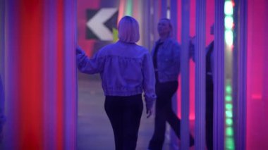 Bir kadın neon ışıklı ayna labirentini keşfeder, duvarlara dokunur ve canlı çevredeki yansımaları gözlemler..