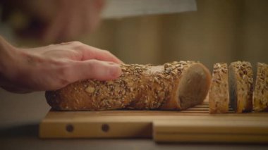 Bir el, mutfak ortamında tahta bir kesme tahtasının üzerinde taze tahıllı ekmek dilimlerine uzanıyor.