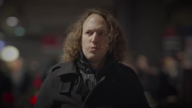 Kıvırcık saçlı bir adam hareketli, bulanık insanlarla çevrilidir ve canlı bir şehir gece manzarasında ışıklar