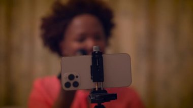 Bir kadın, sıcak bir ortamda modern teknolojiyi gösteren kapalı bir ortamda, tripodun üzerinde akıllı bir telefonla video kaydediyor.