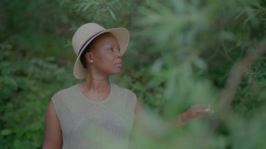 Şapkalı bir kadın, çeşitli bitki yaşamlarıyla çevrili canlı yeşil bir bahçede doğayı keşfeder ve onunla bağlantı kurar.