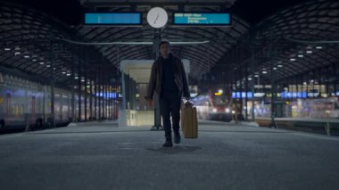 Yalnız bir yolcu, bavuluyla, gece boyunca boş, aydınlık bir tren istasyonunda tek başına bekler.