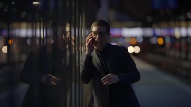 Profesyonel bir iş adamı, gece yarısı şehir ışıklarının cam bir duvara yansıdığı bir telefon görüşmesi yapıyor.