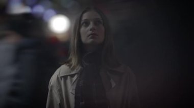 Kışlık ceketli ve atkılı bir kadın, tefekkür eden bir kadın, bir şehir gece sahnesinin arka planına karşı çıkıyor.