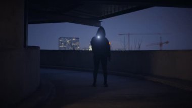 Karanlık bir şehir ortamında elinde bir el feneri olan siluetli bir figür, şehrin arka planına karşı duruyor..