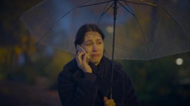 Yağmurlu bir günde parkta bir kadın, şemsiye altında duygusal telefon konuşması yapıyor, yansıtıcı atmosfer yaratıyor.