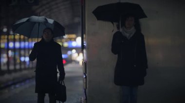 Siyah şemsiyeli iki kişi yağmurlu bir gecede ayrı ayrı şehirde duruyorlar..
