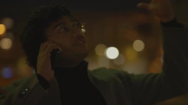 Gözlüklü genç bir adam gece bir telefon görüşmesi yapıyor. Arka planda şehir ışıkları bulanık.