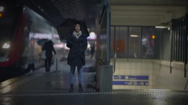 Şemsiyeli kişi yağmurlu bir tren istasyonunda duruyor, gelişini bekliyor, şehir yaşamını yansıtıyor..