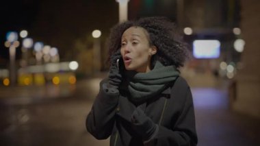 Kentsel bir gece sahnesinde palto ve eşarp giymiş bir kadın, telefonla aranıyor, modern şehir yaşam tarzını yansıtıyor.