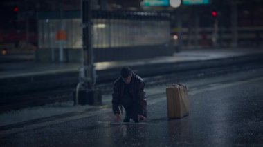 Bavullu yalnız bir yolcu karanlık, yağmurlu bir tren istasyonunda bekler, yalnızlık ve beklenti hisseder.