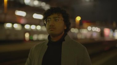 Palto giymiş kıvırcık saçlı genç bir adam, arka planda şehir ışıklarıyla gece bir tren peronunda duruyor..