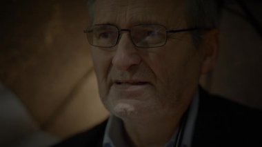 Loş ışıkta, gözlüklü yaşlı bir adam şemsiyenin altında duruyor, düşünceli bir ifade takınıyor.