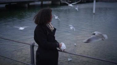 Suyun kenarında kuşları huzur içinde besleyen bir kadın, sakin bir doğa ve yansımayla akşamın tadını çıkarıyor.