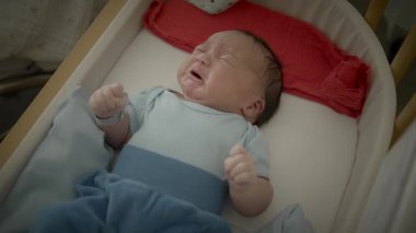 Beşikte ağlayan, mavi tulum giyen bir bebek. Bebek üzgün görünüyor ve beyaz bir çarşafın üzerinde yatıyor..