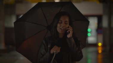 Bir kadın şemsiye tutuyor, gece telefonda konuşuyor, arka planda şehir ışıkları, yağmurlu sokaklar..