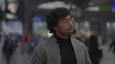 Gözlüklü, kıvırcık saçlı, spor ceket giyen, hareketli bir tren istasyonunda duran düşünceli genç adam..