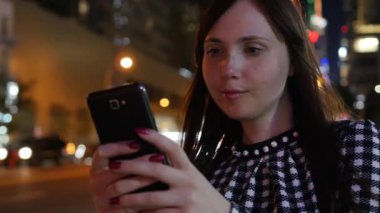 Kareli tişörtlü genç bir kadın gece şehirde arka plan ışıklarıyla aydınlatılmış akıllı telefonunu kullanıyor.